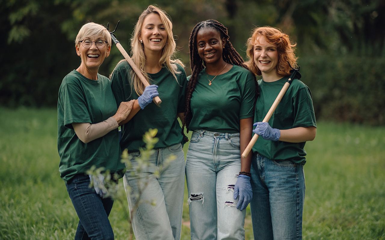 Four female volunteers smiling and holding gardening tools in a park while wearing green shirts